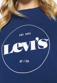 Tmavě modrá košile s bílým logem Levi’s uvnitř kruhu, text „EST. 1873“ nad logem a „SF / CA.“ pod logem.