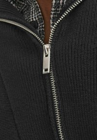 Schwarzer Reißverschluss-Pullover aus strukturiertem Material, mit einem silbernen Reißverschluss und einem sichtbaren karierten Hemdkragen darunter.
