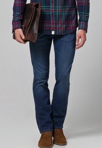 Bolso bandolera de cuero marrón sostenido con una mano, con camisa de cuadros abotonada, jeans azul oscuro y zapatos de ante color beige.