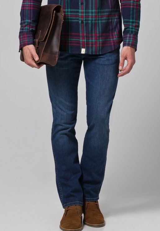 Bolso bandolera de cuero marrón sostenido con una mano, con camisa de cuadros abotonada, jeans azul oscuro y zapatos de ante color beige.