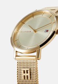 Goldfarbene Armbanduhr mit einem Metallgewebearmband, minimalistischem runden Zifferblatt und subtiler Textur. Verfügt über ein Markenlogo und Stundenmarkierungen.