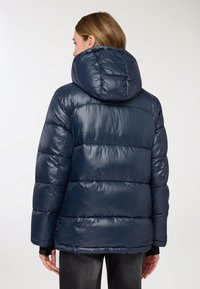 Giacca imbottita blu navy con una texture lucida, dotata di cappuccio e design trapuntato. La giacca ha una vestibilità ampia e polsini elasticizzati.