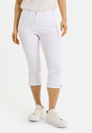 Mujer con pantalones blancos cortos y zapatillas blancas, de pie frente a un fondo claro y liso.