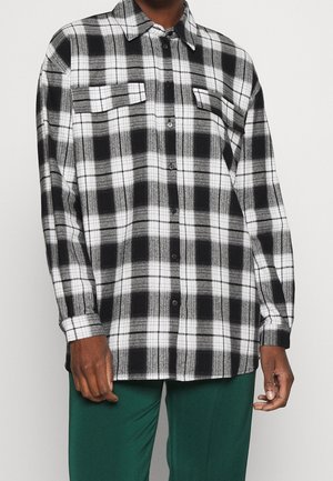 Chemise à carreaux noir et blanc boutonnée avec deux poches poitrine portée par une personne portant un pantalon vert sur un fond uni.