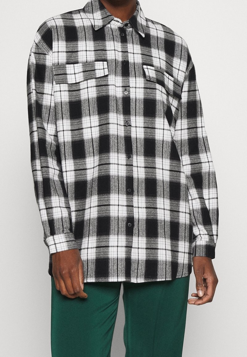 Chemise à carreaux noir et blanc boutonnée avec deux poches poitrine portée par une personne portant un pantalon vert sur un fond uni.