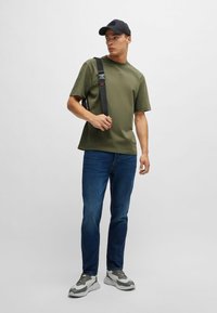 Olivgrön t-shirt, mörkblå jeans, grå och vita sneakers, och en svart keps. Svart bärrem över axeln. Enkelt mönster.