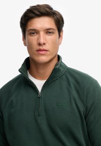 Ciemnozielony pulower z suwakiem do ćwiartki, wykonany z teksturowanej tkaniny, z logo w dopasowanym kolorze na piersi oraz białą wewnętrzną warstwą.