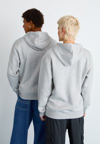 Felpe grigie con una texture morbida, dotate di lacci e tasche a marsupio, indossate da due persone con acconciature diverse.