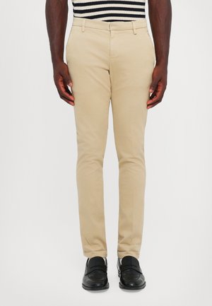 Uomo che indossa pantaloni slim-fit beige, mocassini in pelle nera e una camicia a righe bianco e nera accorciata in vita.
