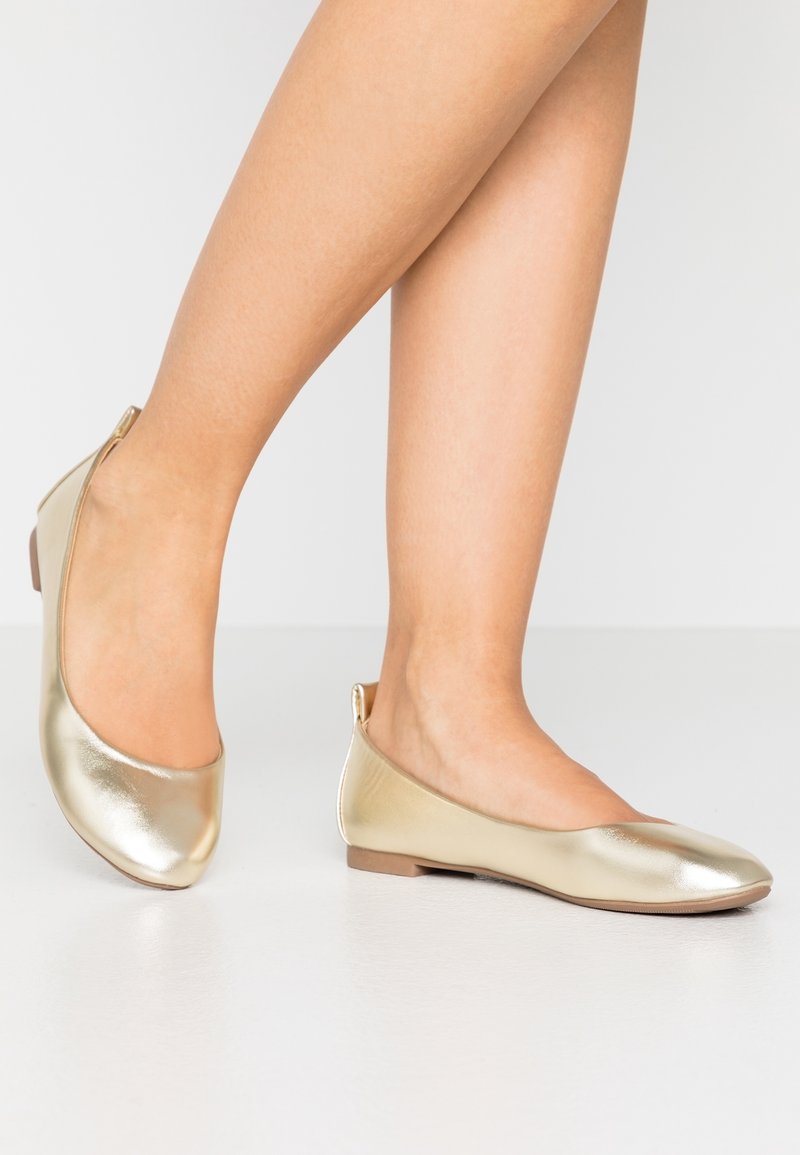 Jambes portant des ballerines dorées brillantes à talons bas sur un fond clair uni.
