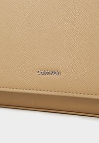 Beżowa torba z teksturowanej skóry marki Calvin Klein z delikatnym przeszyciem i srebrnym logo. Gładkie wykończenie i usztywniony kształt.