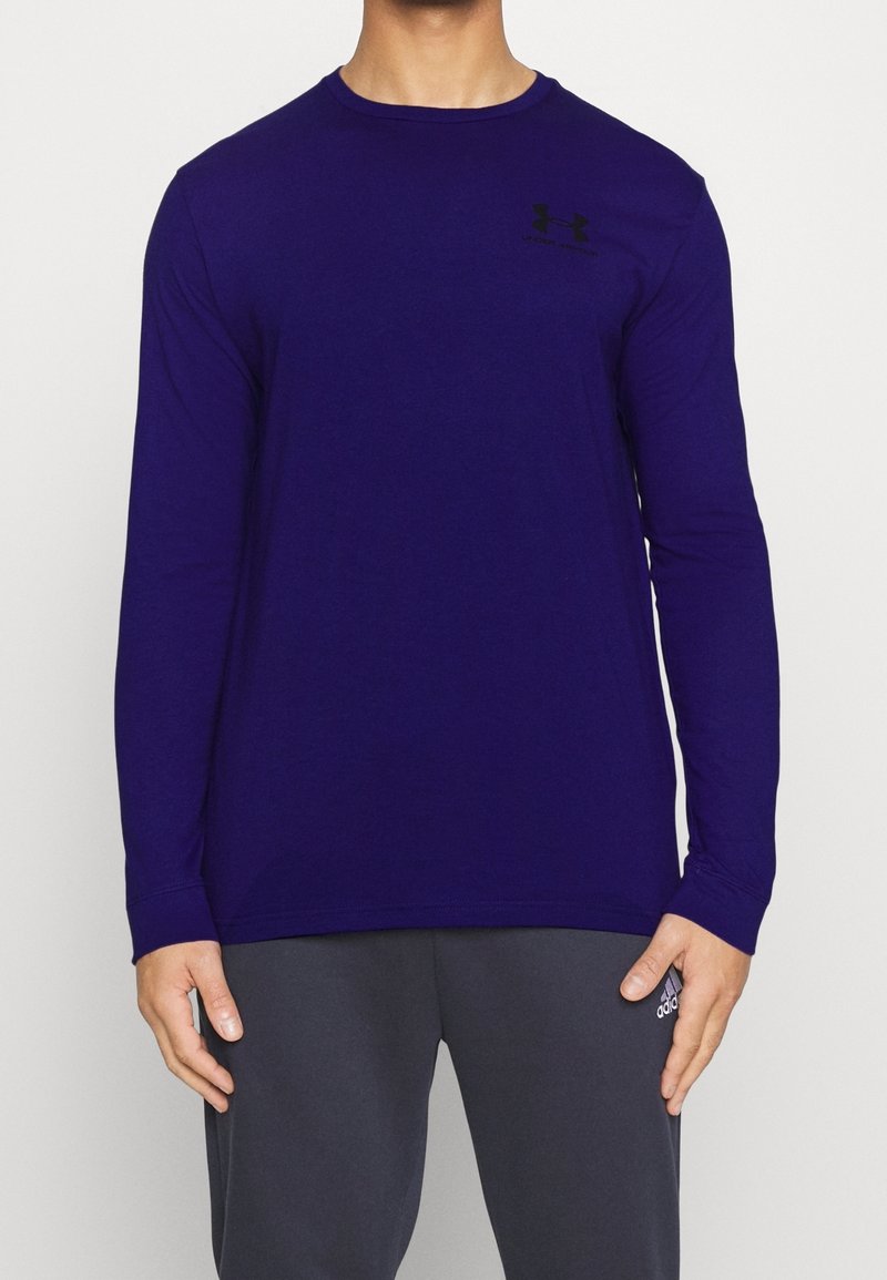 Man som bär en mörkblå långärmad Under Armour-tröja och mörkgrå Adidas byxor står mot en enfärgad bakgrund.