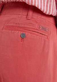 Zblízka na kapsu korálově růžových kalhot s knoflíkem a štítkem značky Polo Ralph Lauren, v kombinaci s červeno-bílou pruhovanou košilí.