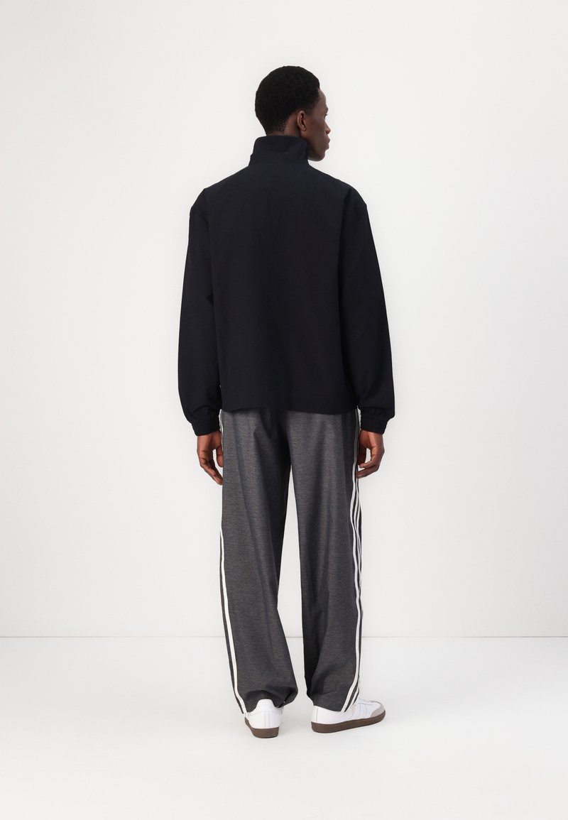 Schwarz hochgeschlossenes Jacket mit langen Ärmeln und grauen Jogginghosen mit weißen Seitenstreifen; glatte, entspannte Passform und lässiges Design.