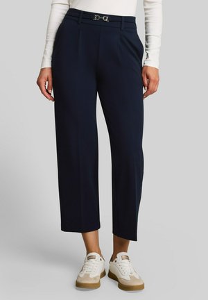 Femme portant un pantalon court bleu marine avec ceinture et un haut blanc à manches longues, assortis à des baskets blanches et beiges sur fond gris.