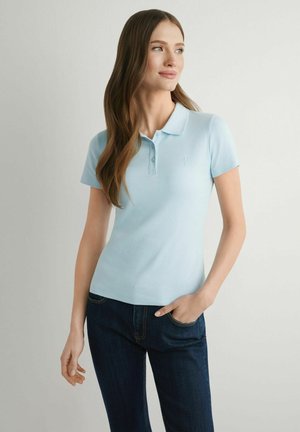 Kobieta z długimi brązowymi włosami, ubrana w jasnoniebieską koszulkę polo z krótkim rękawem i ciemnoniebieskie dżinsy, stojąca z jedną ręką w kieszeni, spoglądająca w prawo.