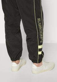 Svarta träningsbyxor med gula detaljer och "LACOSTE"-logotyp på sidan, i kombination med vita sneakers som har en texturerad yta.