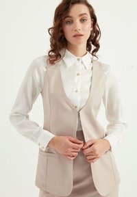 Beige skräddarsydd väst med en slät textur, öppen framsida och två sidofickor, bärs över en vit skjorta med krage och guldknappar.