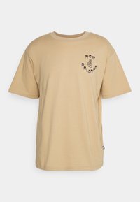 T-shirt en coton beige à manches courtes, avec un logo graphique noir sur la poitrine gauche. Design simple avec un col rond et une coupe régulière.