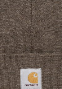 Brązowa dziana tkanina z złożonym szwem i białą kwadratową metką z żółtym logo Carhartt oraz czerwonym napisem.