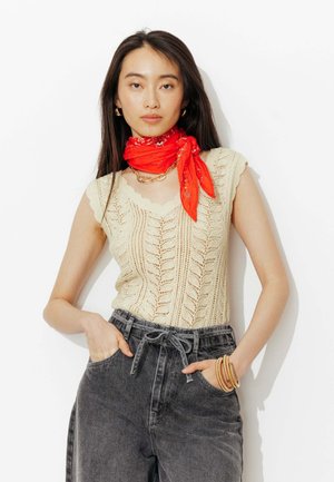 Femme portant un top sans manches en maille crème, un jean noir délavé taille haute, un foulard rouge à motifs autour du cou, un collier chaîne en or, des boucles d'oreilles créoles et des bracelets.