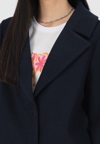 Námořnicky modré sako s širokým klopovým límcem, zapínáním na jeden knoflík a měkkou texturou, vrstvené přes bílé tričko s květinovými vzory.