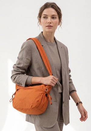 Donna in completo beige con borsa a tracolla arancione con tracolla regolabile, in piedi davanti a uno sfondo chiaro uniforme, che guarda di lato.