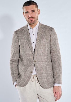 Mann in beigem kariertem Blazer über weißem Hemd mit Knopfleiste und hellen Hose mit Kordelzug, stehend vor einfarbig hellem Hintergrund.