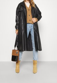 Schwarzer Leder-Trenchcoat mit großen Knöpfen, kombiniert mit einem braunen Strickpullover, hellblauen Jeans und beigen Stiefeletten. Schwarze und braune Handtasche.