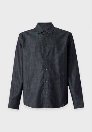 Homme debout les mains dans les poches, portant une chemise sombre à boutons, un pantalon vert olive et des mocassins marron foncé sur un fond uni.