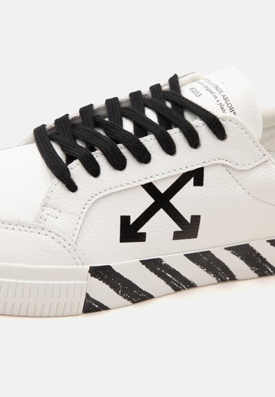 Basket blanche avec lacets noirs, logo flèches croisées noires sur le côté, et rayures diagonales noires sur la semelle.