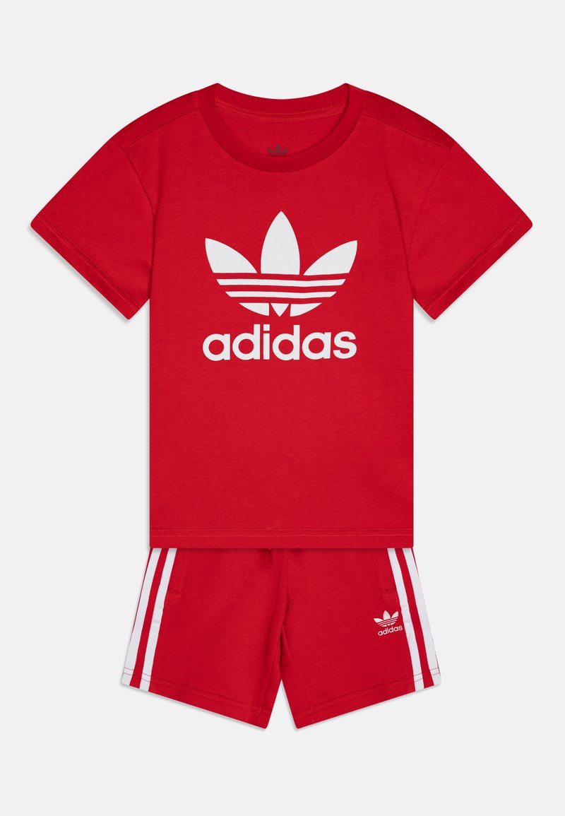 Sarkans kokvilnas sporta komplekts ar t-kreklu, uz kura ir balts adidas logo, un saskaņotām šortiem ar baltiem sānu svītrainēm un logo uz apakšmalas.