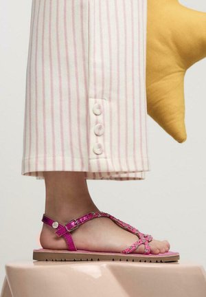Sandalias planas rosas con detalles decorativos en las correas y hebilla, combinadas con pantalones de rayas claras que tienen acentos de botones en el dobladillo.