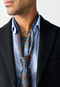 Seidenschal mit komplexem blau-braunem Paisley-Muster, um den Hals gebunden, getragen über einem hellblauen Hemd und einem schwarzen Blazer.