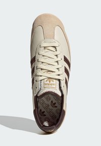 Krémově kožená teniska s hnědými semišovými doplňky, vybavená třemi hnědými pruhy, texturovanými tkaničkami a zlatým logem na vzorované stélce.