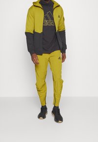 Gelbe Kapuzenjacke mit Reißverschluss und schwarzen Akzenten, kombiniert mit passenden gelben, zulaufenden Hosen. Schwarze Sportschuhe mit strukturierter Oberfläche.