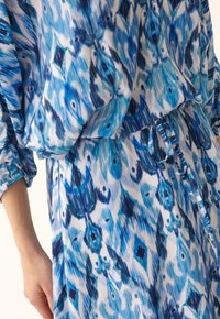 Modrá vzorovaná šaty s volným střihem, obsahující abstraktní design v různých odstínech modré a bílé. Zahrnuje elastické manžety a stahovací pas.