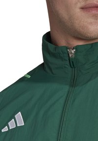 Grüne, leichte Jacke mit einem Reißverschlusskragen, mit einem silbernen Logo auf der Brust und dezenten grünen Akzenten auf den Schultern.