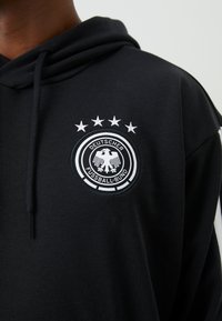 Czarna bluza z kapturem z okrągłą haftowaną naszywką z białymi detalami, pięcioma gwiazdkami powyżej i napisem "DEUTSCHER FUSSBALL-BUND." Materiał bawełniany.