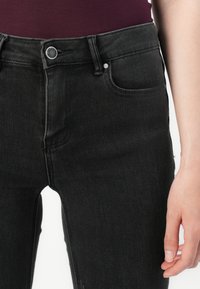 Nærbilde av en person som har på seg svarte denimjeans med detaljer som knapp og lommer foran, og en mørk rød topp som er delvis synlig.