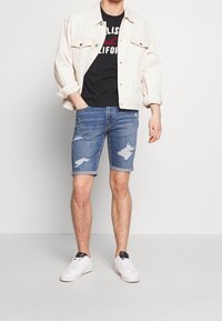 Pantalones cortos de mezclilla con parches desgastados, una camiseta negra clásica y una chaqueta de mezclilla crema. Zapatillas blancas completan el atuendo casual.