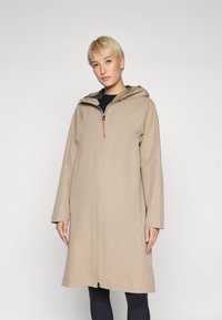 Beige vesihylkivä takki, jossa on vetoketjullinen etuosa, huppu ja pitkät hihat. Sisältää korostetun punaisen vetoketjun vedin ja löysän, suoran leikkauksen.