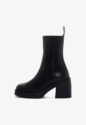 Botte de cheville en cuir noir avec un talon bloc et une languette à l'arrière, finition lisse et bout arrondi, photographiée sur fond blanc.