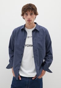 Camicia a quadri blu con maniche lunghe sopra una maglietta bianca con grafica. Jeans in denim slim fit. Modello in piedi contro uno sfondo semplice.
