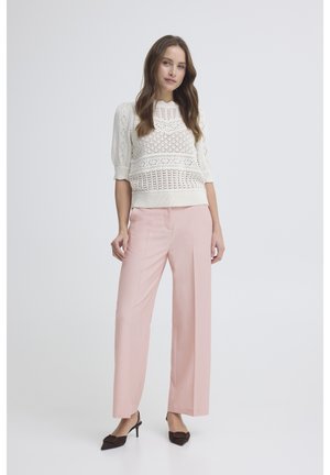 Nő áll, fehér kötött rövid ujjú pulóvert, világos rózsaszín bő szárú nadrágot és sötétbarna hegyes orrú magas sarkút viselve, egyszínű háttér előtt.