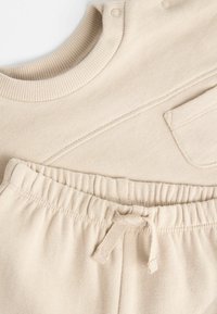 bézs, két részes szett, amely ribbed crewneck topból áll gombokkal, valamint egy párba illő, elasztikus derékpánttal ellátott nadrágból, amely húzózsinórral és oldalsó zsebbel rendelkezik.