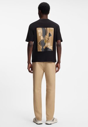 Homme portant un T-shirt noir avec un texte graphique beige "Wine & Music" dans le dos, un pantalon beige et des baskets blanches, debout de dos.