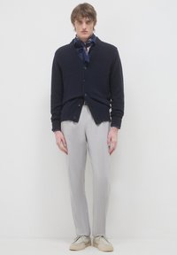 Giovane uomo con un cardigan blu navy sopra una camicia scura e una sciarpa al collo, pantaloni grigio chiaro e scarpe beige, in piedi su uno sfondo semplice.