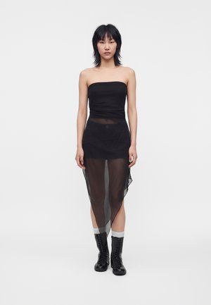 Vrouw met kort zwart haar die een strapless zwarte jurk draagt met een doorschijnende asymmetrische rok en zwarte veterschoenen met witte sokken.
