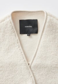 Indumento in pile bianco con superficie testurizzata, caratterizzato da una scollatura a V profonda e un'etichetta nera con la scritta "someday." L'etichetta della taglia indica "36."